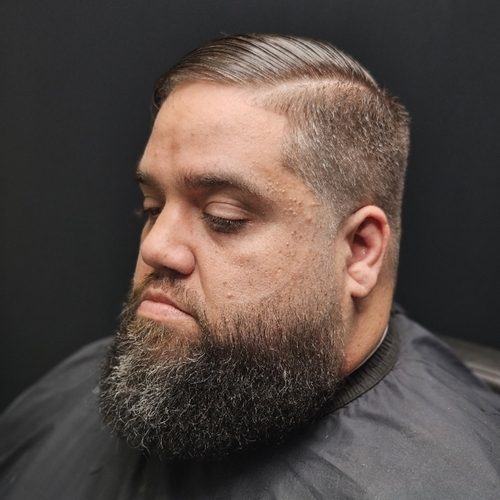 Man with a slicked-back side part, tapered sides and a full beard shaped and lined